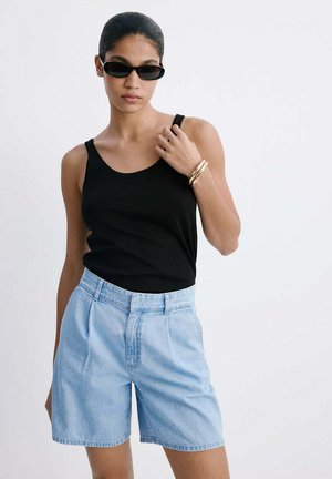 Femme portant un haut noir sans manches, un short en jean taille haute bleu clair, des lunettes de soleil noires et des bracelets dorés sur fond blanc.