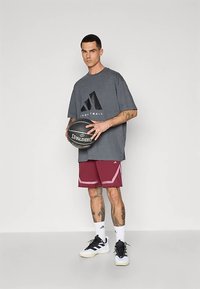 Camiseta gris de gran tamaño con gráfico negro de baloncesto, pantalones cortos color burdeos con detalles en blanco, zapatillas deportivas blancas y negras, sosteniendo un balón de baloncesto.