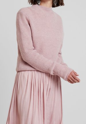 Femme portant un pull en maille duveteuse rose pâle associé à une jupe plissée rose pâle et des boucles d'oreilles géométriques en argent.