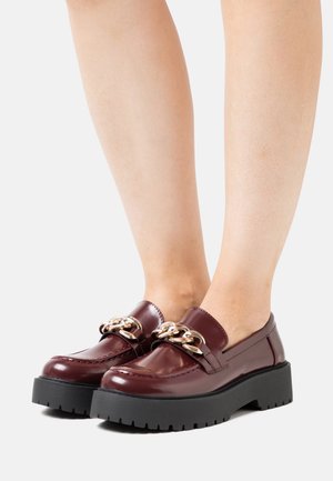 Mocassins en vernis bourgogne avec détail en chaîne dorée, semelle plateforme noire texturée et accents cousus, présentant un design élégant et moderne.