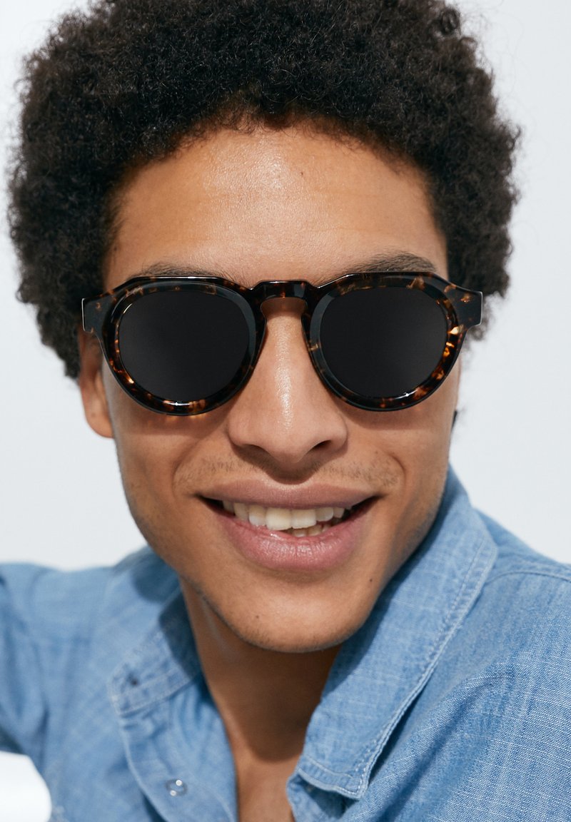 Gafas de sol tortuga redondas con lentes oscuros. Montura texturizada y forma redonda prominente. Las lleva puestas una persona con cabello rizado y una camisa de mezclilla.