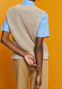 Personne portant un gilet en tricot beige par-dessus une chemise bleu à manches courtes et un pantalon beige, se tenant contre un fond orange avec les mains derrière le dos.