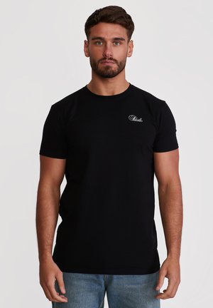 Muž s krátkými hnědými vlasy a vousy, oblečený v jednoduchém černém tričku s malým bílým logem "SikSilk" na hrudi, stojící před bílým pozadím.