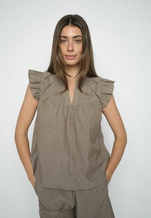 Hosszú barna hajú nő, laza, ujjatlan, fodros vállú taupe színű felsőt és hozzá illő nadrágot visel, kezeit a zsebében tartva áll.