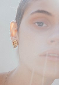 Kuldne kõrvarõngas tekstureeritud ringikujulise disainiga ja väikese riputusega aksessooriga, kantud vasakus kõrvus, pehmelt uduses taustal.