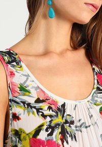 Top sin mangas de flores con escote cuadrado que presenta patrones en rosa, verde y amarillo; combinado con pendientes azul en forma de gota.