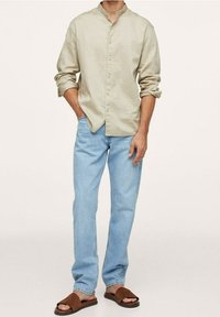 Jasno beżowa koszula z przyciskiem i kołnierzykiem mandaryńskim, w połączeniu z jasnoniebieskimi dżinsami i brązowymi klapkami; casualowy i luźny fason.