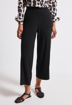 Pantalons noirs, court, à large jambe en tissu doux et extensible. Taille élastique pour le confort. Associés à des sandales tressées noires.