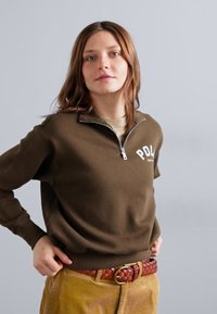 Καφέ φούτερ με φερμουάρ και λευκό λογότυπο, συνδυασμένο με πλεχτή κόκκινη ζώνη. Το μοντέλο φοράει παντελόνια σουέτ, αναδεικνύοντας υφές και επίπεδα.