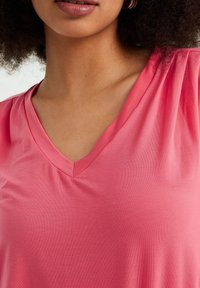 Tričko s výstřihem do V v jasně růžové barvě, vyrobené z hladké, lehké látky. Detailní záběr ukazuje jemnou strukturu a subtilní lesk.