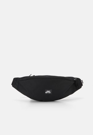 Sac banane Nike SB noir avec poche avant zippée et sangle ajustable sur fond gris clair.