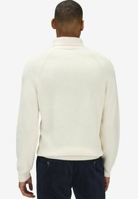 Ribbed white sweater with a high collar and raglan sleeves, seen from the back, paired with dark textured corduroy pants.