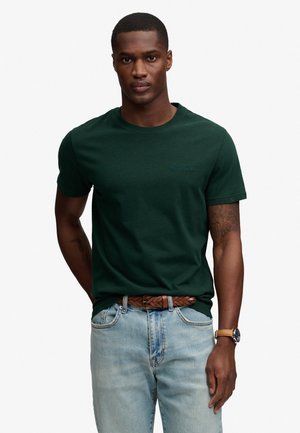 Mężczyzna ubrany w ciemnozieloną koszulkę z krótkim rękawem, jasnoniebieskie dżinsy z plecionym brązowym paskiem oraz zegarek na rękę, stojący na białym tle.