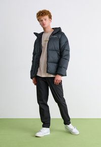 Schwarze Pufferjacke mit Kapuze, gesteppte Textur, Reißverschluss vorne, Logo auf der Brust. Tragen über einem beige T-Shirt und schwarzen Hosen mit weißen Sneakers.