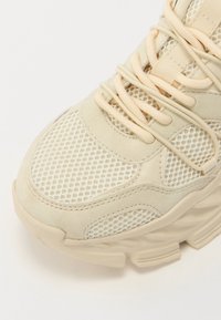 Beige athletic shoe with mesh and suede material. Rounded shape features prominent lacing and textured accents on the upper.