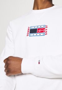 Vit sweatshirttopp med en färgglad broderad "TOMMY JEANS"-logotyp, med svarta, blå och röda accenter. Ärmarna har en liten trikolor-logodetalj.