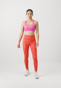Reggiseno sportivo rosa e leggings con un vivace motivo geometrico in arancione e giallo. Modella che indossa scarpe da ginnastica bianche, in piedi contro uno sfondo neutro.
