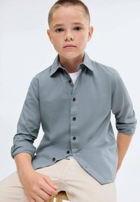 Poika, jolla on lyhyet hiukset, istuu tuolilla harmaassa nappipaidassa, jonka hihat on käännetty ylöspäin, ja vaaleissa housuissa valkoista taustaa vasten.