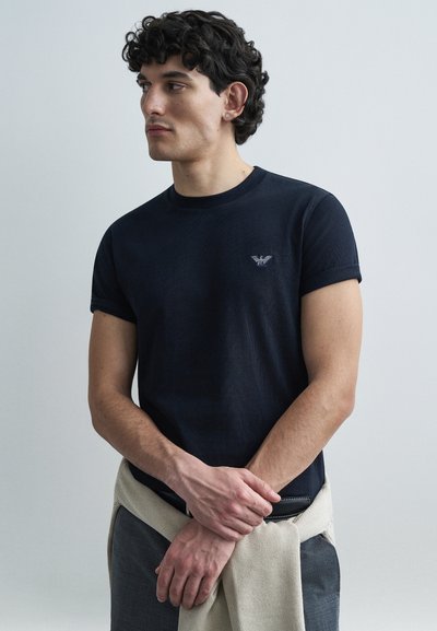 Marineblå T-shirt med korte ærmer og ribbet struktur, med en lille broderet logo på brystet. Brugt sammen med en lys trøje bundet om livet.