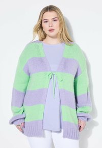 Strickjacke mit grünen und lavendelfarbenen Streifen, offenes Vorderteil mit einer Bindeschließe, weite Ärmel und strukturierten Stoff. Trägt man über einem lavendelfarbenen Oberteil.