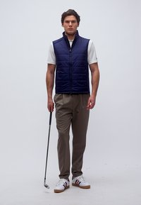 Άνδρας με μπλε γιλέκο, λευκή πόλο και μπεζ παντελόνι, κρατώντας ένα γκολφ ρόπαλο, στέκεται μπροστά σε απλό ανοιχτόχρωμο φόντο.