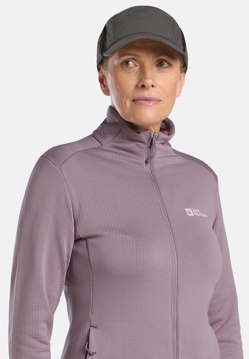 Veste zippée violette avec col montant, tissu texturé, poches latérales et logo Jack Wolfskin sur la poitrine, associée à une casquette noire.