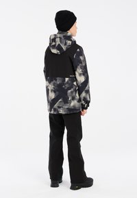 Schwarz-graue gemusterte Jacke mit Kapuze, die ein schwarzes Panel an der Taille aufweist. Kombiniert mit schwarzen Hosen und robusten schwarzen Stiefeln.