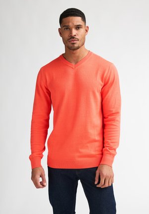 Mann mit kurzen Haaren, der einen leuchtend orangefarbenen V-Ausschnittpullover und dunkelblaue Jeans trägt, steht vor einem schlichten hellen Hintergrund.