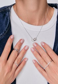 Ręce prezentujące kilka pierścionków oraz naszyjnik z połączonymi okręgami, na tle białej bluzki i dżinsowej kurtki.