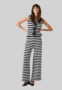 Zwart-wit gestreepte jumpsuit met wijde pijpen, gestructureerde stof en vetersluiting aan de voorkant. Gecombineerd met zwarte schoenen.