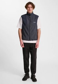 Mörkblå ärmlös fleecväst med dragkedja framtill och ståkrage, kombinerad med en vit T-shirt, svarta byxor och mörka sneakers.