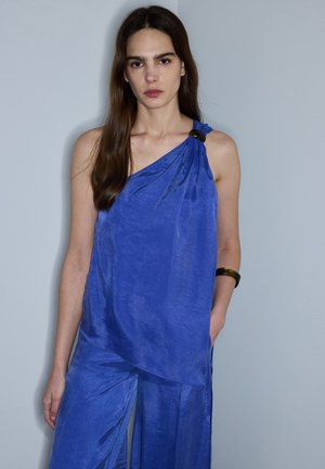 Vrouw met lang bruin haar draagt een mouwloos koningsblauw one-shoulder topje en bijpassende wijde broek tegen een grijze achtergrond.