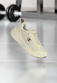 Αθλητικό παπούτσι σε κρεμ απόχρωση με χοντρή σόλα και λογότυπο Champion, αιωρείται πάνω από έναν ανακλαστικό πάτωμα γυμναστηρίου με βάρη στο φόντο.