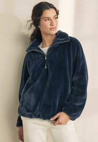 Pulover din fleece bleumarin, cu guler înalt, fermoar pe jumătate în față și manșete elastice. Textură moale și croială lejeră. Purtați peste un top ușor.