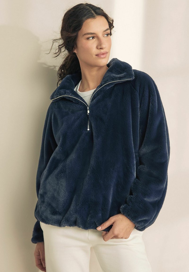 Pulover din fleece bleumarin, cu guler înalt, fermoar pe jumătate în față și manșete elastice. Textură moale și croială lejeră. Purtați peste un top ușor.
