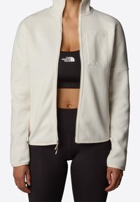 Kobieta w rozpiętej kremowej polarowej kurtce nosi czarny stanik sportowy i legginsy, obie rzeczy z logo The North Face na środku klatki piersiowej.