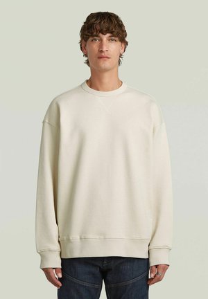 Giovane uomo con capelli ricci che indossa una felpa crema semplice e oversize e jeans scuri, in piedi su uno sfondo chiaro.