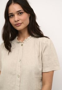 Beige linned blouse med korte ærmer, rynket krave og tre knapper fortil, med en subtil tekstur.