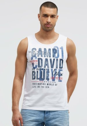 Man wearing white sleeveless shirt with blue and red text about sea life and a pair of blue jeans, standing against plain background.