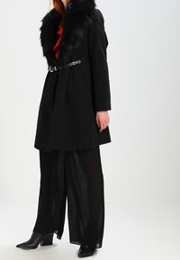 Svart kappa med krage av fuskpäls, bälte i midjan, matchad med genomskinliga svarta plisserade byxor och stövletter med klack. Enkel, elegant design.