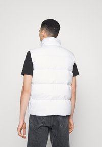 Vit puffertill med hög krage, med kviltade sektioner. Bärs över en svart skjorta, kombineras med mörkgrå jeans. Enkelt design, utan utsmyckningar.