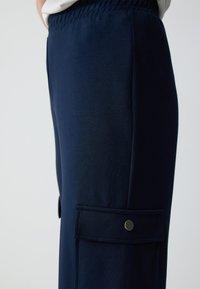 Donkerblauwe broek van gladde stof met een zijzak met een metalen drukknoopsluiting. Elastische tailleband, taps toelopend design.