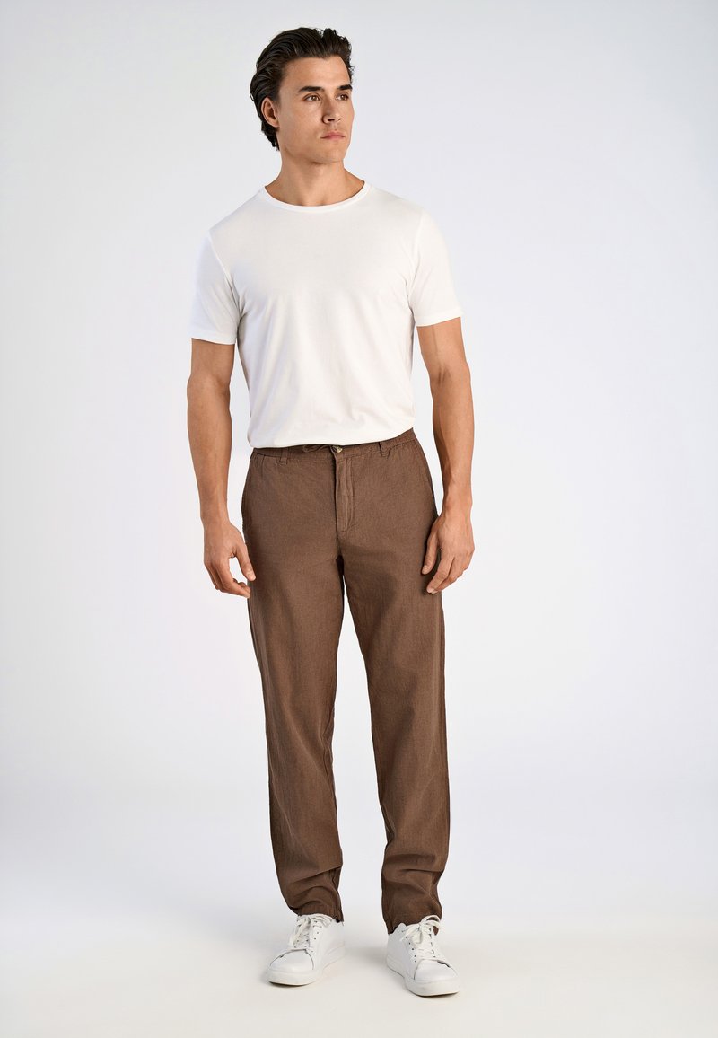 Witte t-shirt met korte mouwen gecombineerd met bruine linnen broek met een trekkoord in de taille, rechte pijpen en witte sneakers.