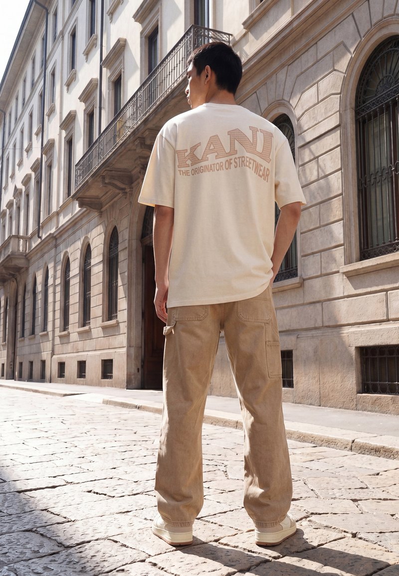 Mann steht auf einer Kopfsteinpflasterstraße, trägt ein beiges KANI-T-Shirt und beige Cargohosen und blickt auf ein steinernes Gebäude mit bogenförmigen Fenstern.