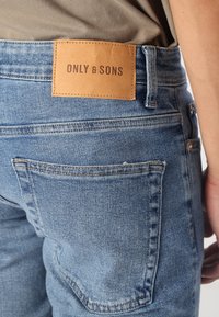 Jeansy jeansowe w jasnoniebieskim kolorze o gładkiej fakturze, z brązową skórzaną naszywką z logo "ONLY & SONS" na tylnej części pasa.