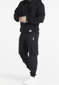 Person iført sort hættetrøje og sorte cargo bukser med lapper, hænderne foldet, og hvide sneakers med guld detaljer, stående foran en hvid baggrund.