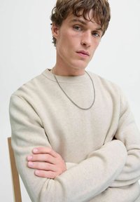 Cremefarbener Pullover mit geripptem Ausschnitt und strukturiertem Stoff, mit langen Ärmeln. Eine silberne Kettenhalskette hängt vorne herab.