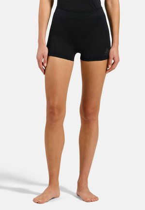 Femme portant un short de sport noir moulant, debout pieds nus devant un fond clair uni.