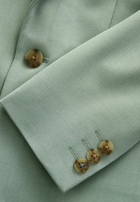 Tissu vert clair avec une manche ajustée ornée de trois boutons marron et d'accents en chaîne, mettant en avant une texture lisse et un design structuré.