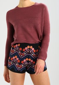 Mörkröd långärmad topp med texturerad stickad design, kombinerad med svarta shorts med färgglada zigzag-mönster och blommig brodyr.